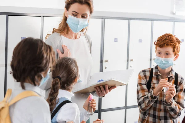 Pupils holding hand sanitizer near teacher in medical mask with notebook and infrared thermometer — Stock Photo