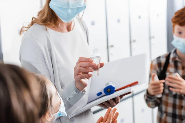 Teacher with notebook and infrared thermometer holding hand sanitizer near pupils in medical masks — Stock Photo