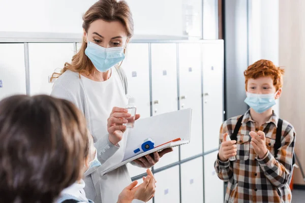 Teacher with disinfector, infrared thermometer and notebook standing near kids in corridor — Stock Photo