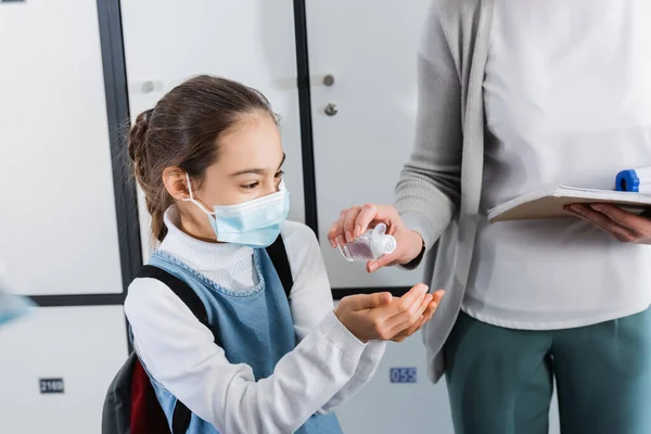 Teacher with infrared thermometer and notebook pouring sanitizer on hands of pupil in medical mask — Stock Photo