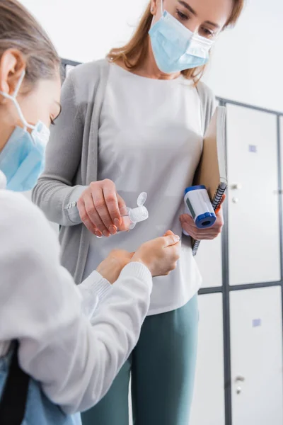 Disinfector in hand of teacher with infrared thermometer and notebook near schoolgirl on blurred foreground — Stock Photo