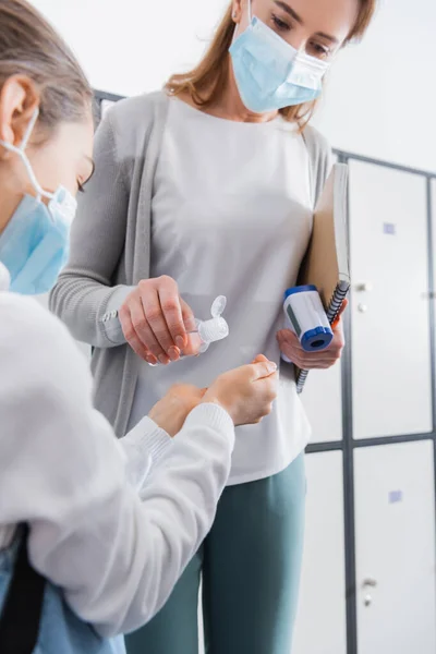 Desinfektionsmittel in der Hand eines Lehrers mit Infrarot-Thermometer und Notizbuch in der Nähe von Schülerin im unscharfen Vordergrund — Stockfoto