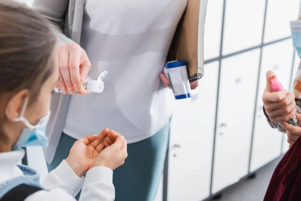 Profesor con termómetro infrarrojo y portátil vertiendo desinfectante en las manos de la pupila en máscara médica - foto de stock