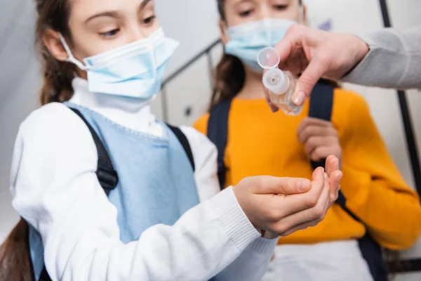 Insegnante versando disinfettante mano sulle mani di allievo in maschera medica su sfondo sfocato — Foto stock
