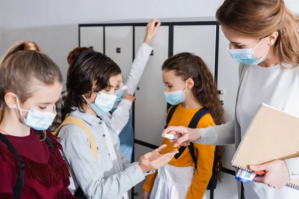 Lehrer mit Notizbuch und Infrarot-Thermometer hält Desinfektionsmittel in der Nähe der Hände von Schülern — Stockfoto