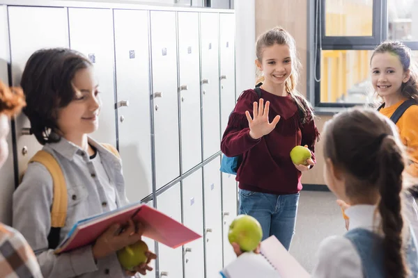 Smiling schoolgirl with apple waving hand to classmates in school corridor — Stock Photo