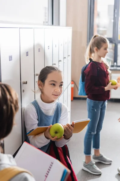 Colegiala hablando con amigos y sosteniendo cuaderno y manzana en la sala de la escuela - foto de stock