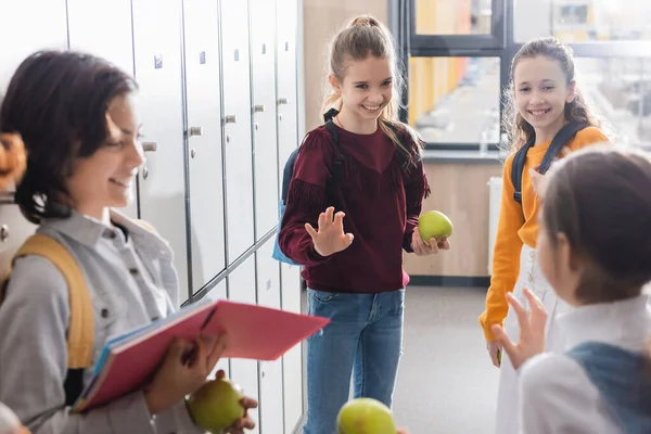 Colegiala con manzana y mochila saludando mano a amigos en primer plano borroso en la sala de la escuela - foto de stock