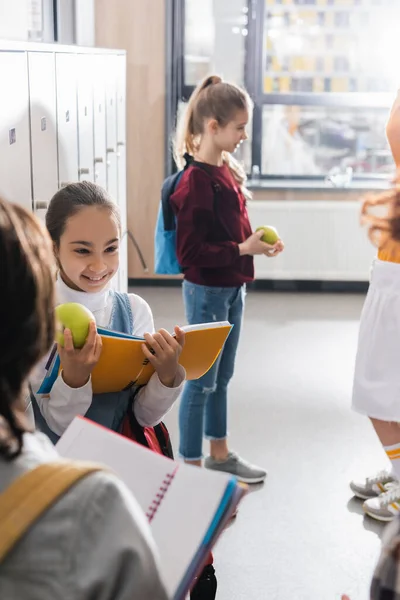 Веселая школьница с яблоком и блокнотом, смотрящая на одноклассников на размытом переднем плане в холле — стоковое фото
