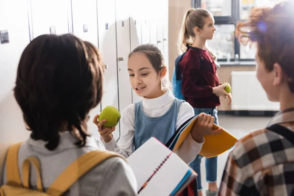 Colegial con cuaderno mirando manzana cerca de colegiales en primer plano borroso en el pasillo - foto de stock