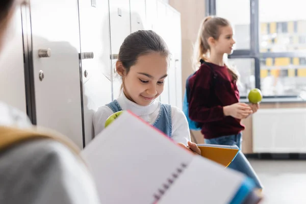 Niño sonriente con manzana mirando el cuaderno cerca de su compañero de clase en primer plano borroso en la escuela - foto de stock
