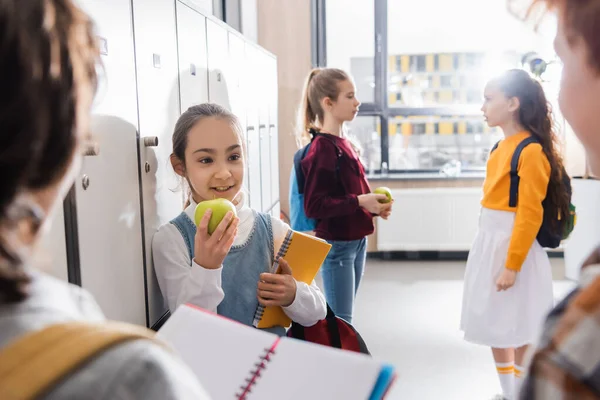 Niño feliz sosteniendo manzana madura y cuaderno mientras mira a los escolares en primer plano borroso - foto de stock