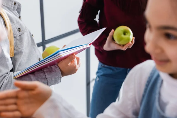 Quaderno e mela nelle mani dello scolaro vicino agli amici in primo piano sfocato — Foto stock