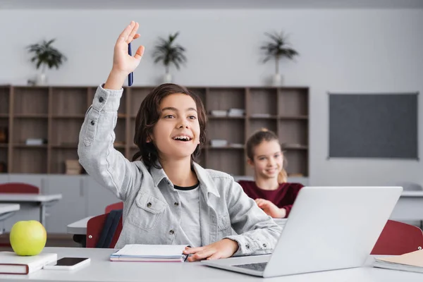 Cheerful boy raising hand near notebook and devices in classroom — Stock Photo