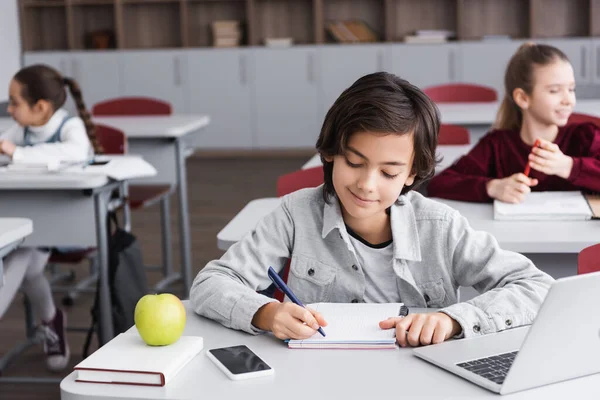 Schoolkid writing on notebook near gadgets, book and apple — Stock Photo