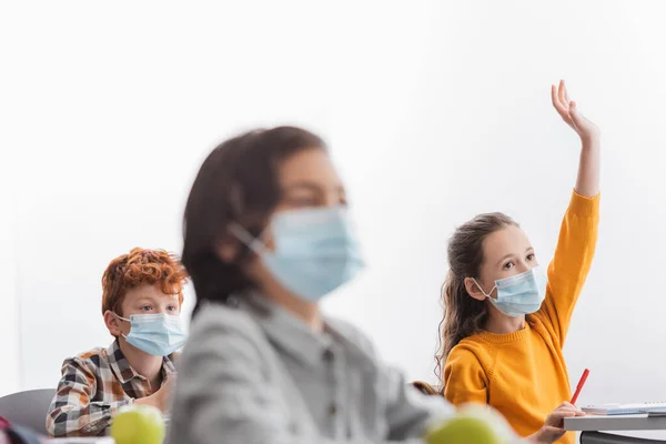 Schoolgirl in medical mask raising hand near classmates in class — Stock Photo