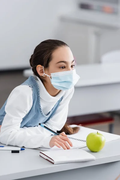 Chica en máscara protectora sentado cerca de portátil y manzana en el escritorio en la escuela - foto de stock