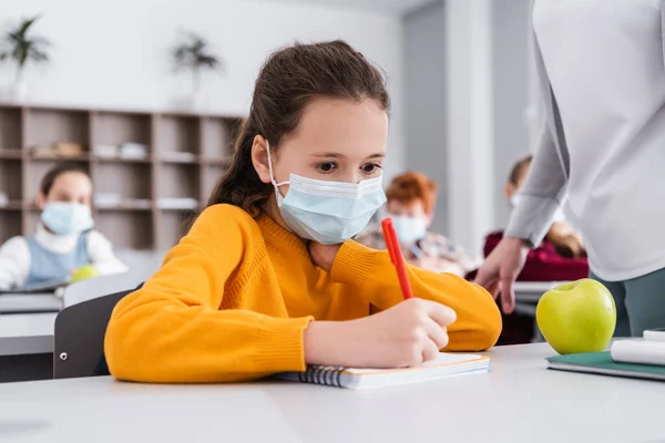 Schoolgirl in medical mask writing in notebook near teacher and schoolmates on blurred background — Stock Photo