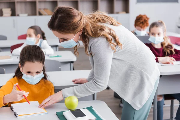 Teacher in medical mask looking at notebook near schoolgirl during lesson — Stock Photo