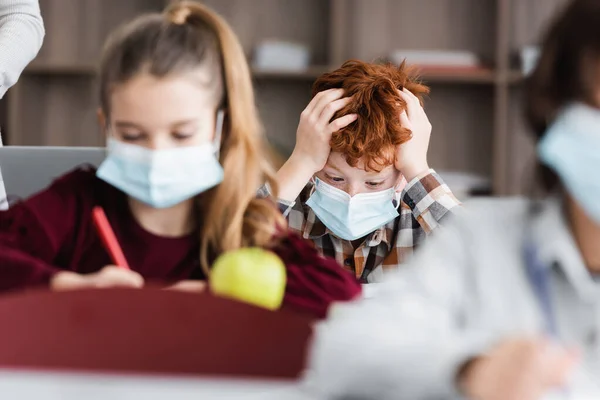 Tired, redhead schoolkid in medical mask touching head near classmates on blurred foreground — Stock Photo