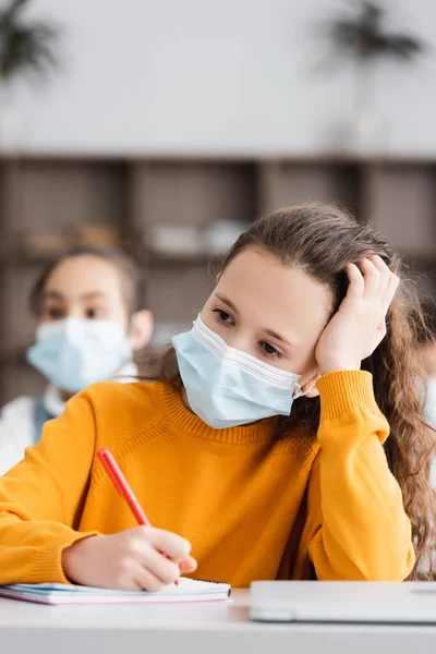 Pensive schoolkid in medical mask writing in notebook in classroom — Stock Photo