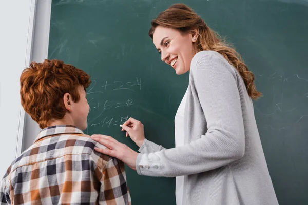 Happy teacher touching shoulder of redhead schoolboy near chalkboard — Stock Photo