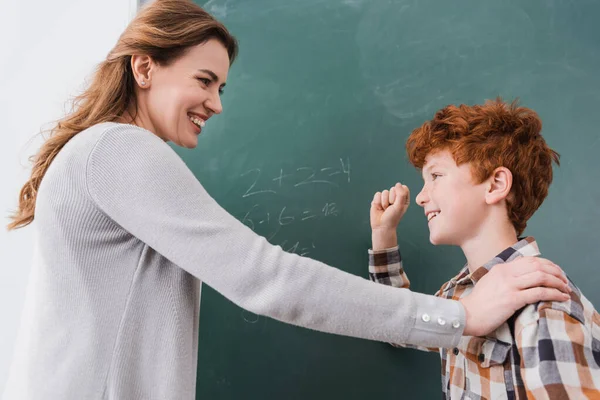 Smiling teacher touching shoulder of schoolboy showing win gesture near chalkboard — Stock Photo