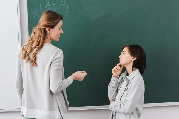 Pensativo colegial mirando sonriente profesor cerca de pizarra - foto de stock