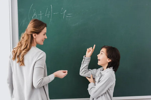 Colegial excitado apuntando con el dedo a pizarra cerca de profesor sonriente - foto de stock