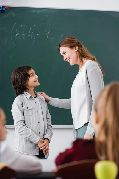 Cheerful schoolboy and teacher looking at each other near chalkboard and pupils on blurred foreground — Stock Photo