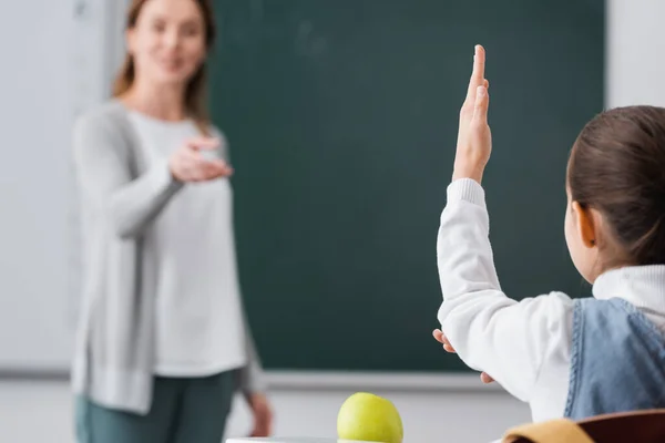 Back view of schoolgirl raising hand near teacher on blurred background — Stock Photo