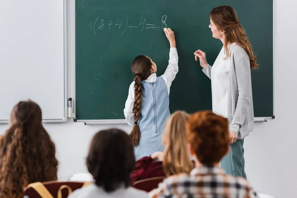 Enseignant près de écolière écriture sur tableau, et les élèves sur flou au premier plan — Photo de stock