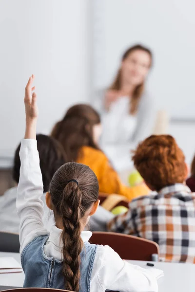 Back view of schoolgirl with raised hand near classmates and teacher on blurred background — Stock Photo