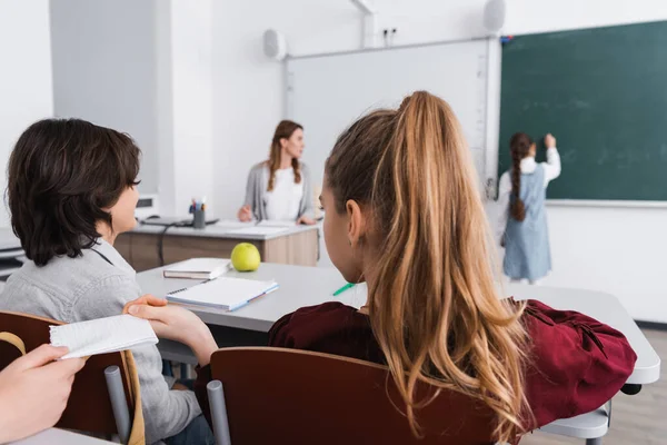 Schoolgirl taking note from classmate near pupils and teacher on blurred background — Stock Photo