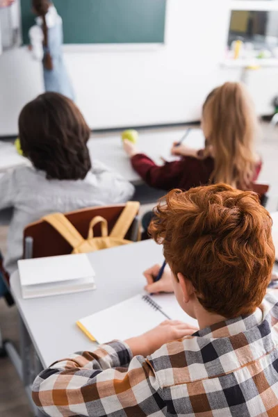 Back view of pupils writing in notebooks in classroom, blurred background — Stock Photo