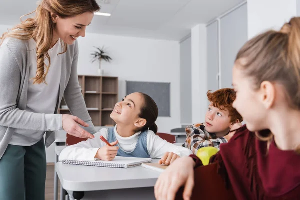 Happy teacher and schoolgirl looking at each other in classroom near pupils on blurred foreground — Stock Photo