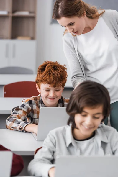Insegnante in piedi vicino sorridente rossa ragazzo utilizzando computer portatile durante la lezione — Foto stock