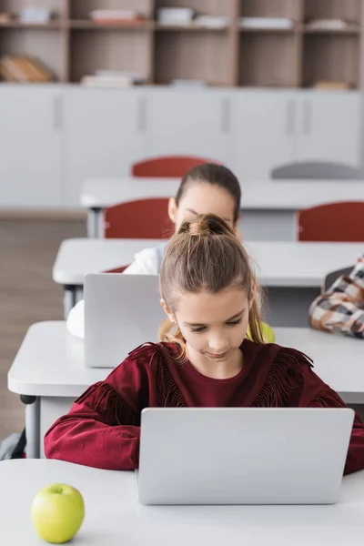 Chica usando el ordenador portátil durante la lección cerca de compañeros de clase en fondo borroso - foto de stock