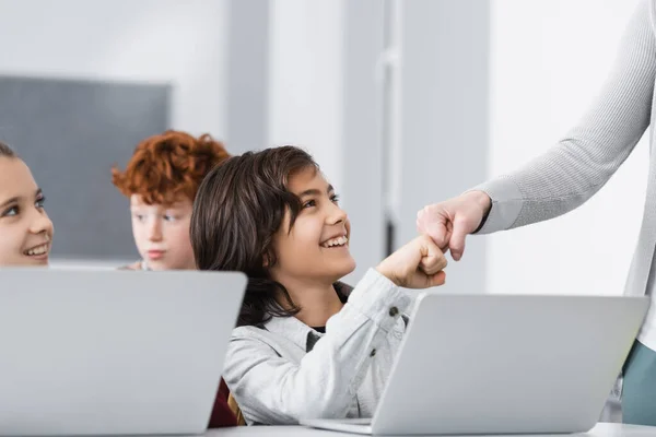 Веселый школьник делает удар кулаком с учителем рядом с ноутбуком и одноклассниками на размытом фоне — стоковое фото