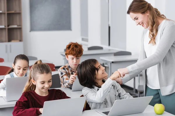 Profesor emocionado y colegial haciendo golpe de puño cerca de la computadora portátil y los alumnos en el aula - foto de stock