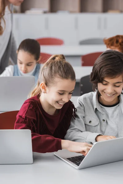 Excited kids typing on laptop near classmates on blurred background — Stock Photo