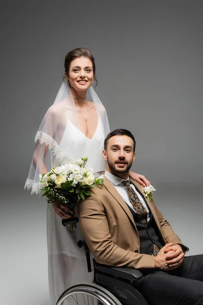 Bride with bouquet and arabian groom in wheelchair smiling at camera isolated on grey — Stock Photo
