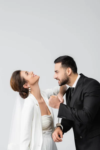 Wütender muslimischer Bräutigam in Handschellen blickt fröhliche Braut isoliert auf grau an — Stockfoto