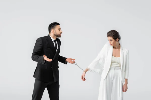 Muslim groom showing shrug gesture near bride in handcuffs isolated on grey — Stock Photo