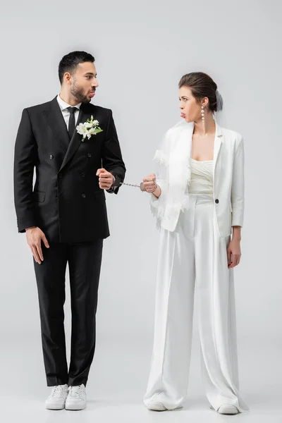Multikultureller Bräutigam und Braut in Handschellen, die die Zunge auf weißem Hintergrund herausstrecken — Stockfoto