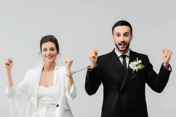 Cheerful interracial couple in wedding clothes and handcuffs showing yes gesture isolated on grey — Stock Photo