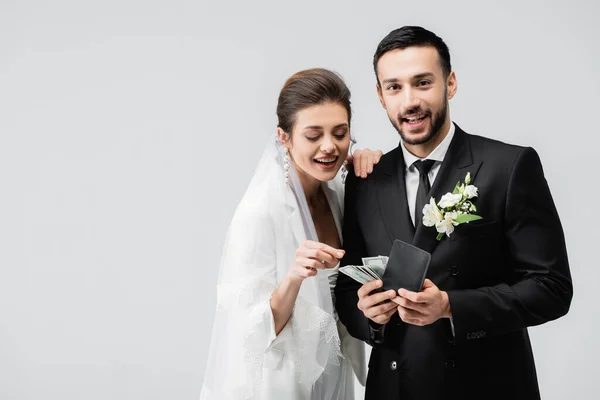 Novia sonriente mirando a la billetera con dinero en manos del novio musulmán aislado en gris - foto de stock