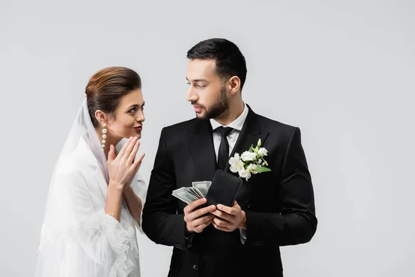 Novia alegre mirando novio árabe con billetera y dinero aislado en gris - foto de stock