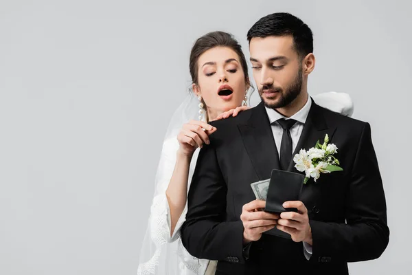 Noiva excitada olhando para carteira com dinheiro em mãos de noivo muçulmano isolado em cinza — Fotografia de Stock