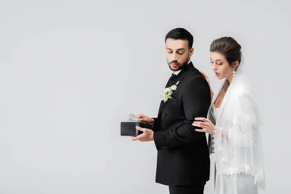 Mariée excitée debout près du marié arabe avec de l'argent et portefeuille isolé sur gris — Photo de stock